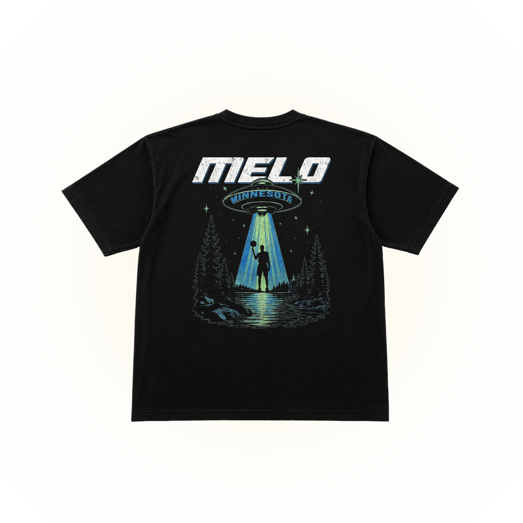 The back side of a black comfort colors tshirt featuring a large graphic print on the back. The graphic displays the text 'MELO MINNESOTA' in white and blue, above an illustration of a UFO sending a beam of light to a silhouetted figure standing in a forest by a lake.