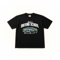 A black short-sleeve comfort colors graphic t-shirt featuring the text 'Melo's DRIVING SCHOOL' in white above a stylized green and blue car illustration, displayed flat against a solid white background.